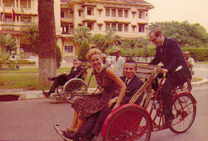 Audrey Topping, author and photojournalist, rides in a Cambodian cyclo pedicab, with her husband, Seymour Topping, then chief Southeast Asia correspondent for The New York Times. In this photo taken in 1963, Eric Pace, Times correspondent in Saigon pedals in front of Phnom Penh's Hotel Le Royal, while his Times colleague from Saigon, Jack Langguth, watches.