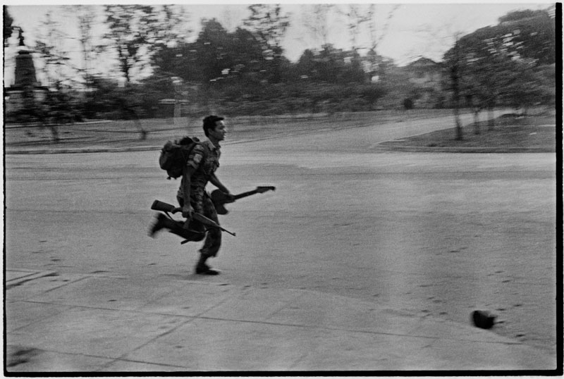 A Khmer Republic soldier runs toward the center of Phnom Penh with his guitar and rifle at dawn on April 17, 1975, as government forces fight the Khmer Rouge on the outskirts of the city. (Roland Neveu)