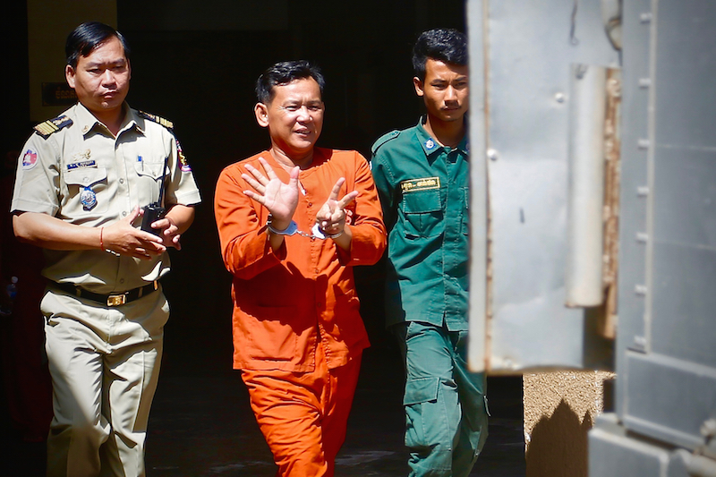 Seang Chet is escorted from the Phnom Penh Municipal Court on Monday after being sentenced to five years in prison over bribery charges relating to CNRP Vice President Kem Sokha's 'prostitution' case. He is holding up seven fingers to represent the CNRP's position on the 2013 national election ballot. (Siv Channa/The Cambodia Daily)